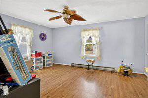 A room with light purple walls, two windows with sheer curtains, wood flooring, a ceiling fan with leaf-shaped blades, some storage bins, a stool, a heater, and various household items in the corners.