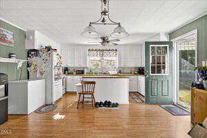 A kitchen with white cabinets, a stove, and a refrigerator covered in magnets and papers. A wooden chair is at the counter, shoes are lined up on the floor, and a green door is open to a backyard. Natural light fills the room.