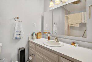 A bathroom with a white countertop, single sink, faucet, mirror, towel rack with a plastic bag, and various toiletries. A towel hangs over the shower door in the background. The walls are painted light gray.