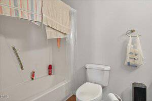 A bathroom with a white bathtub and shower, towels hanging on the shower rod, two bottles on the tub edge, a white toilet, a plastic bag hanging on a hook, and a small dark trash bin on the floor.