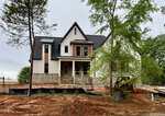 A large two-story house under construction with stone and siding exterior, front porch, and steep gable roof. Construction equipment and dirt are in the front yard, with trees and temporary barriers around the site.