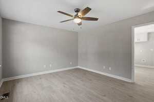 Empty room with light wood flooring, light gray walls, a ceiling fan with wooden blades and a light, and an open doorway leading to another room with similar flooring and partial view of white cabinets.