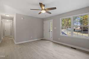A bright, empty room with light wood flooring, beige walls, a ceiling fan with wooden blades, a white entrance door, and a large window showing a view of trees and a street outside.