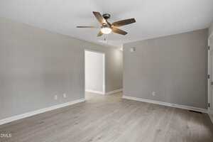 A clean, empty room with light wood flooring, beige walls, a ceiling fan with lights, and an open doorway leading to another area. There are multiple electrical outlets on the walls.