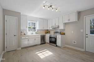 A modern kitchen with white cabinets, stainless steel appliances, light wood flooring, a window above the sink, and a door to the outside. The room is well-lit with ceiling spotlights and natural light.