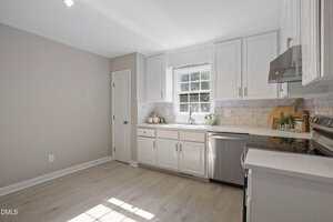 A modern kitchen with white cabinets, light wood flooring, stainless steel appliances, a window above the sink, and neutral-colored walls. A cutting board, utensils, and small plants are on the countertop.