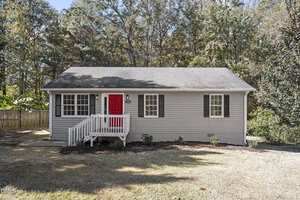 Nestled at 318 Apple Street in Creedmoor, this small, single-story house features light gray siding, black shutters, and a charming red front door. A white porch railing and a few small shrubs complete the inviting look. Trees and a wooden fence sit behind.