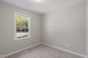 An empty room with beige carpet, light gray walls, a white-framed window letting in natural light, and electrical outlets visible on the right wall. A vent is located on the floor beneath the window.