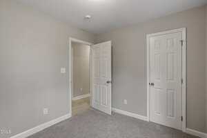 A small, empty room with light gray walls, beige carpet, white baseboards, two white doors, and a ceiling-mounted smoke detector. One door is slightly open, leading to a hallway with light wood flooring.