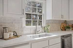 A bright kitchen with white cabinets and countertop, a stainless steel sink and faucet, light gray tile backsplash, and decorative items including cutting boards, a pitcher, greenery, and a "Welcome" sign. Sunlight comes through the window.