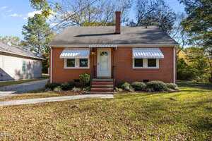 A small brick house at 300 Coleman Street in Oxford, featuring a central white door, two windows with white awnings, a chimney, and a short staircase. The front yard has grass, shrubs, and a paved walkway under trees and blue sky.