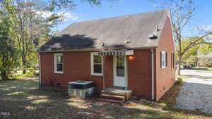 A small red brick house with a steep roof, two windows, and a white back door with a metal awning. An HVAC unit sits by the door, and there are trees, fallen leaves, and a gravel driveway visible.
