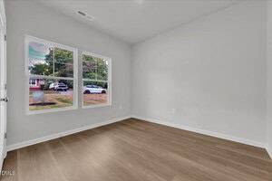 A minimally furnished room with light-colored walls, wood flooring, two large windows showing a street view with trees and cars outside, and a white baseboard trim. No furniture is present in the room.