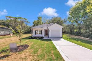 Single-story house with white siding, a covered front porch, and a brown front door. There is a one-car garage, a concrete driveway, a small lawn, and trees in the background under a blue sky.