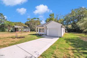 A small, single-story white house with a gray roof and attached garage sits on a grassy lot with a concrete driveway. Trees and another house are visible in the background under a blue sky with scattered clouds.