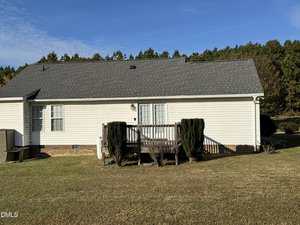 A small wooden porch with steps and trimmed shrubs in front of a one-story white house with gray shingles, brick foundation, and a grassy yard. Trees are visible in the background under a blue sky.