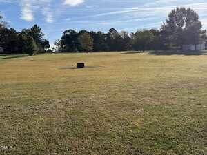 A large, grassy field with a circular concrete structure in the center. Trees border the background under a blue sky with scattered clouds. Shadows from the trees fall across parts of the field.