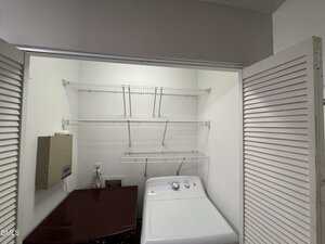 A laundry area with a white washing machine, a dark-colored dryer, three wire shelves mounted on the wall, an electrical panel, and folding louvered doors partially open.