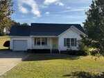 1318 Bobbitt Road in Kittrell is a single-story white house with a covered front porch, attached one-car garage, and dark roof. It sits on a grassy yard with trees and a driveway, beneath a partly cloudy sky.