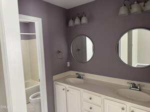 A bathroom with two sinks, two circular mirrors, white cabinetry, and a purple wall. A door leads to another bathroom area with a bathtub and a toilet. Three-light fixtures are mounted above the mirrors.