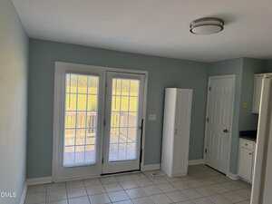 A room with light blue walls, white tile floor, double glass doors with blinds leading outside, a tall white cabinet, a door to the right, and a ceiling light fixture.
