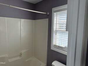 A bathroom with a cream-colored shower enclosure, a silver shower curtain rod, a window with white blinds, and a partial view of a white toilet under the window. The walls are painted a light grayish purple color.
