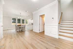 A bright, modern open-concept dining and kitchen area with light wood floors, white walls, a dining table with chairs, recessed lighting, and a staircase leading upstairs.