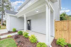 Front view of a modern house with light gray siding, a covered entryway, black door with a wreath, small shrubs along the walkway, and a wooden privacy fence to the right. Blue sky with scattered clouds in the background.