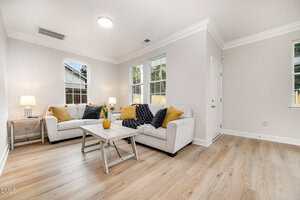 A bright living room with light wood flooring features two beige sofas with yellow and black cushions, a white coffee table, two lamps, and several windows letting in natural light.