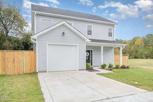 A two-story light gray house with white trim, a single-car garage, covered front porch, wooden fence on each side, and a concrete driveway, set against a grassy yard and trees under a partly cloudy sky.