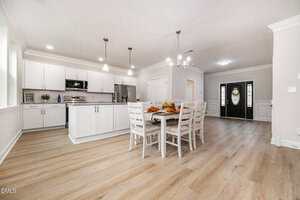 Open-concept kitchen and dining area with white cabinets, stainless steel appliances, a wooden dining table with chairs, pendant lights, light wood flooring, and a view of a black front door.