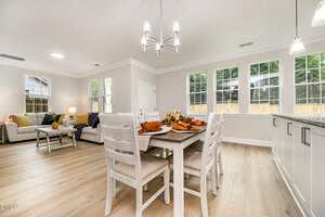 A bright, modern open-concept living and dining area with light wood flooring, white walls, large windows, and a dining table set for four. The living room has sofas and a coffee table near the windows.
