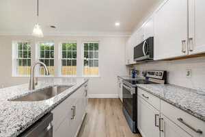 A modern kitchen with white cabinets, granite countertops, stainless steel appliances, a center island with a sink, pendant lighting, and large windows letting in natural light.