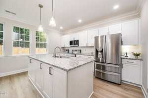 Modern kitchen with white cabinets, stainless steel appliances, granite countertops, a center island with a sink, pendant lighting, light wood flooring, and large windows letting in natural light.