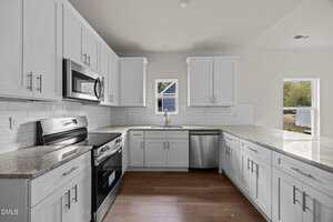 A modern kitchen with white cabinets, stainless steel appliances, granite countertops, white subway tile backsplash, wood flooring, and two small windows above the sink letting in natural light.