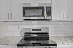 A stainless steel microwave is mounted above a matching electric stove with a smooth black cooktop. The kitchen features white cabinets, white subway tile backsplash, and a speckled light granite countertop.