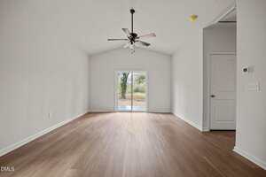 A spacious, empty room with light wood flooring, white walls, a ceiling fan with lights, and a sliding glass door leading outside. A closed white door and a wall switch are visible on the right wall.
