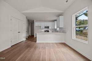 A modern kitchen with white cabinets, stainless steel appliances, and a center island is shown next to an open area with light wood flooring and a large window overlooking a yard with trees.