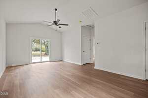A bright, empty room with light wood flooring, white walls, a ceiling fan, and sliding glass doors leading outside. There are two closed white doors and a small hallway visible on the right.