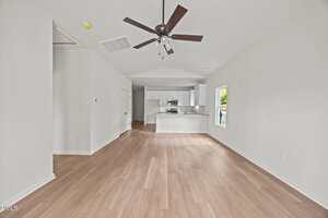 A modern, open-concept living space with light wood flooring, white walls, a ceiling fan, and a view into a white kitchen with stainless steel appliances and a window letting in natural light.