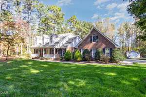 A two-story suburban house at 6018 W Tom Parham Road, Oxford, features red brick and gray siding, surrounded by trees and shrubs, with a large front yard and a detached garage in the background under a blue sky.
