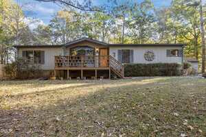 A single-story house on 1644 Fawn Lane in Creedmoor features light gray siding, a wooden front porch and steps, and a large decorative wreath. Surrounded by trees, grass, and fallen leaves, it offers cozy charm in every season.