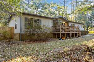 A single-story house with light-colored siding and brick foundation features a wooden front porch with railings, surrounded by trees and fallen leaves on the ground.