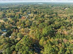Aerial view of a rural, wooded area with scattered houses and roads surrounded by dense green and autumn-colored trees under a partly cloudy sky.