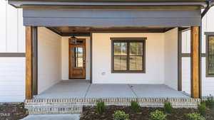 A covered front porch with wood posts, a wooden front door with glass panels, a single window, white siding, and a small landscaped area with shrubs in front of a brick-lined concrete porch.