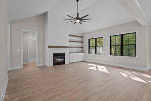 A bright, empty living room with light wood flooring, white walls, black-framed windows, a modern ceiling fan, a white shiplap fireplace, and built-in wooden shelves and cabinets. Sunlight streams in through the windows.