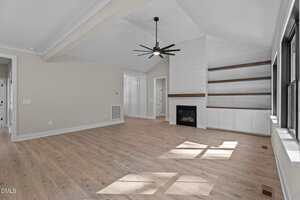 A modern, empty living room with light wood floors, beige walls, a black ceiling fan, a gas fireplace, built-in white shelves and cabinets, and large windows letting in natural light.