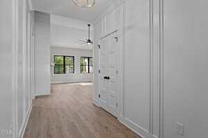 A hallway with light wood flooring and white paneled walls leads to a bright living area with large black-framed windows, a ceiling fan, and modern light fixtures. Double doors are on the right side of the hallway.