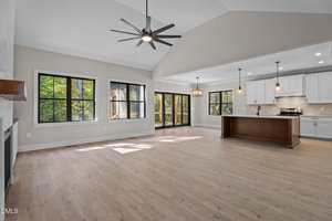 A spacious, empty open-plan living area with light wood flooring, large windows, a fireplace, a central kitchen island, white cabinetry, modern pendant lights, and sliding glass doors to the outside.