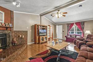 A cozy living room with a brick fireplace, a wooden entertainment center with a TV, a red sofa set, a coffee table with framed photos, hardwood floors, and red curtains covering a window.